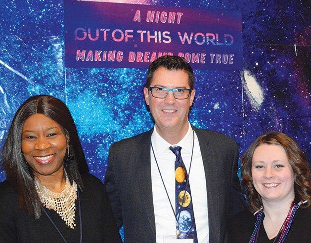Three people pose for a photo in front of a banner that reads "A Night Out of This World, Making Dreams Come True"